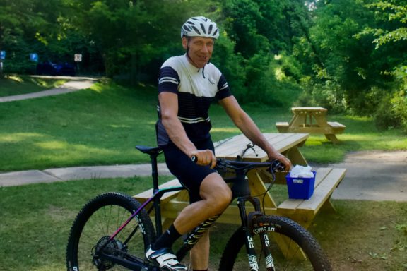 A cyclist in casual attire sits on a bench near a picnic area in a park.