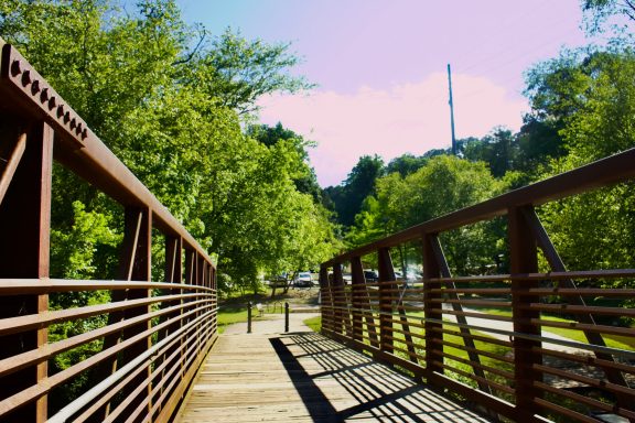 Metal bridge leading through lush green trees under a clear sky.